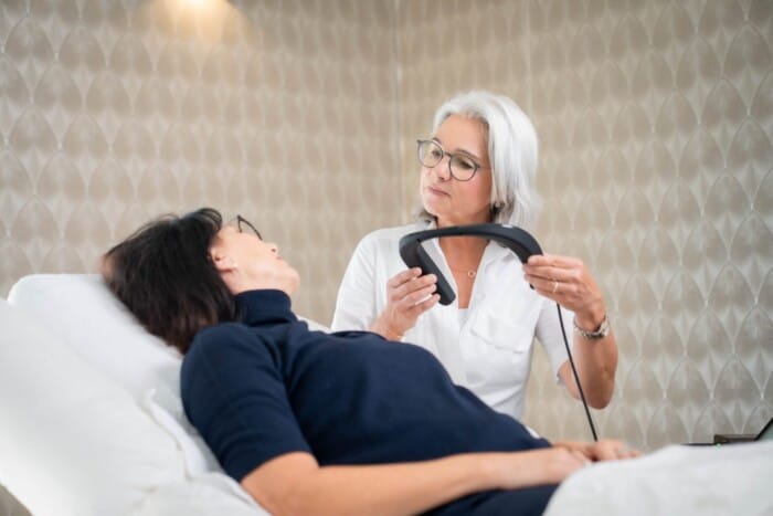 Die Kundin liegt entspannt auf einer Liege, die Kosmetikerin hält das Messgerät, um es der Kundin um den Nacken zu platzieren.