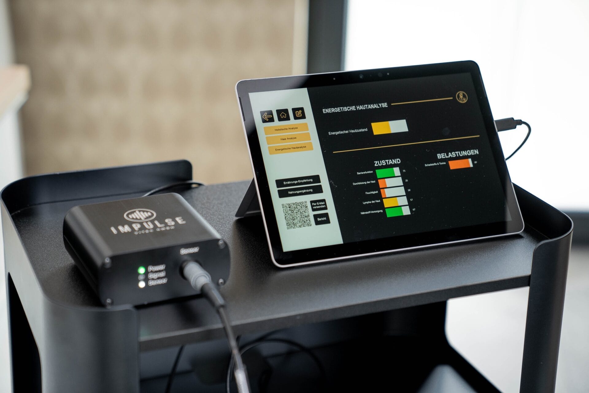 Das kleine Impulse Messgerät und daneben steht das Tablet, auf dem einige Messergebnisse zum energetischen Hautzustand zu sehen sind.