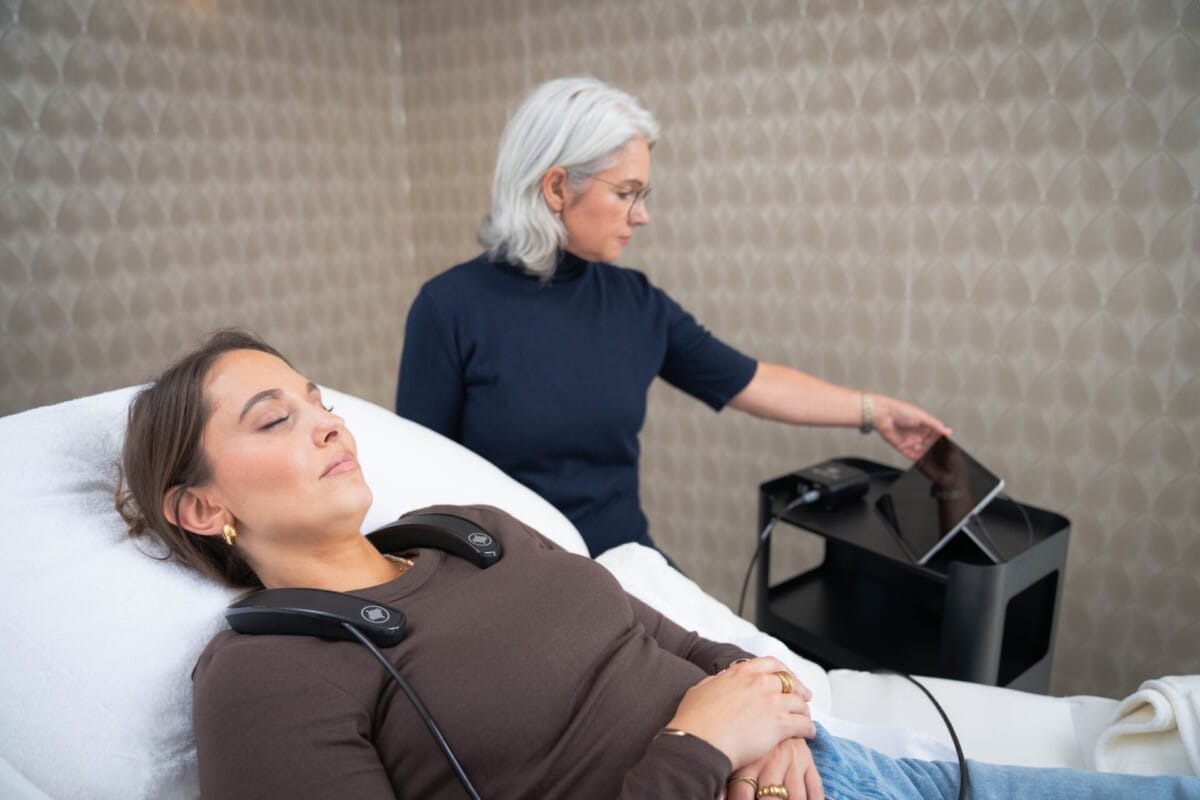 Im Vordergrund liegt entspannt mit geschlossenen Augen die Kundin mit dem Messgerät um den Nacken, im Hintergrund überwacht die Kosmetikerin die Messung