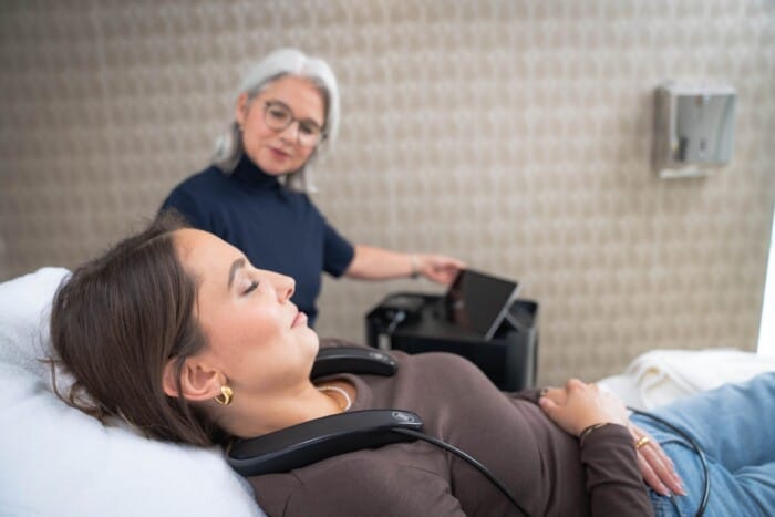 Im Vordergrund liegt entspannt mit geschlossenen Augen die Kundin mit dem Messgerät um den Nacken, im Hintergrund überwacht die Kosmetikerin die Messung