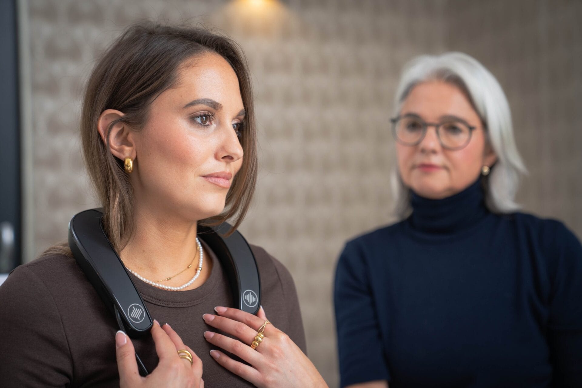 Im Vordergrund eine Kundin mit dem kleinen, leichten Messgerät um den Nacken gelegt. Im Hintergrund leicht unscharf die Kosmetikerin.