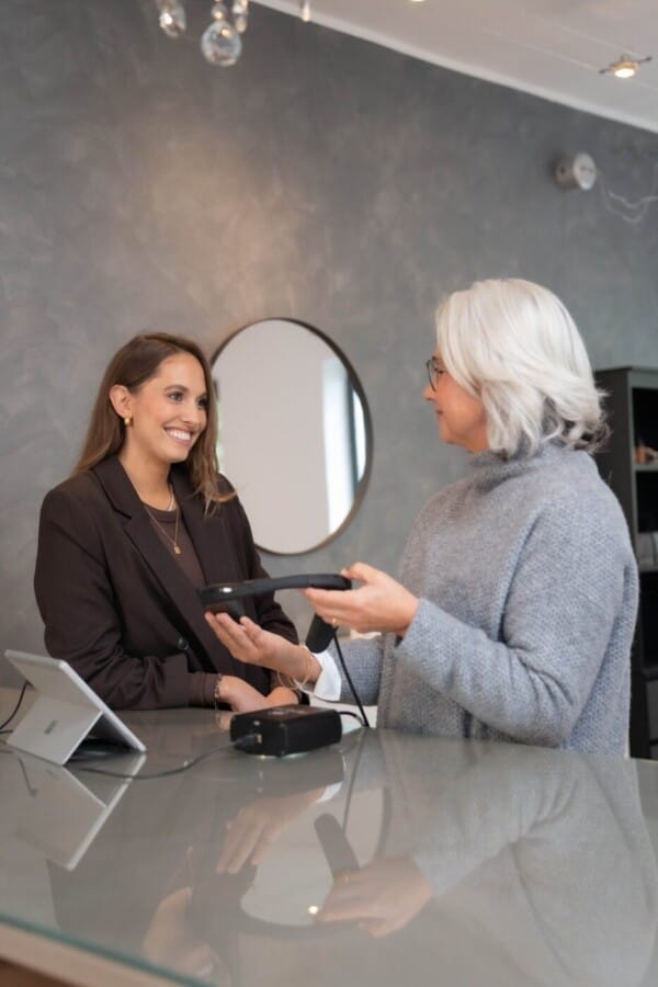 Kosmetikerin hält das Beauty-Check Messgerät in der Hand und präsentiert es der lächelnden Kundin.